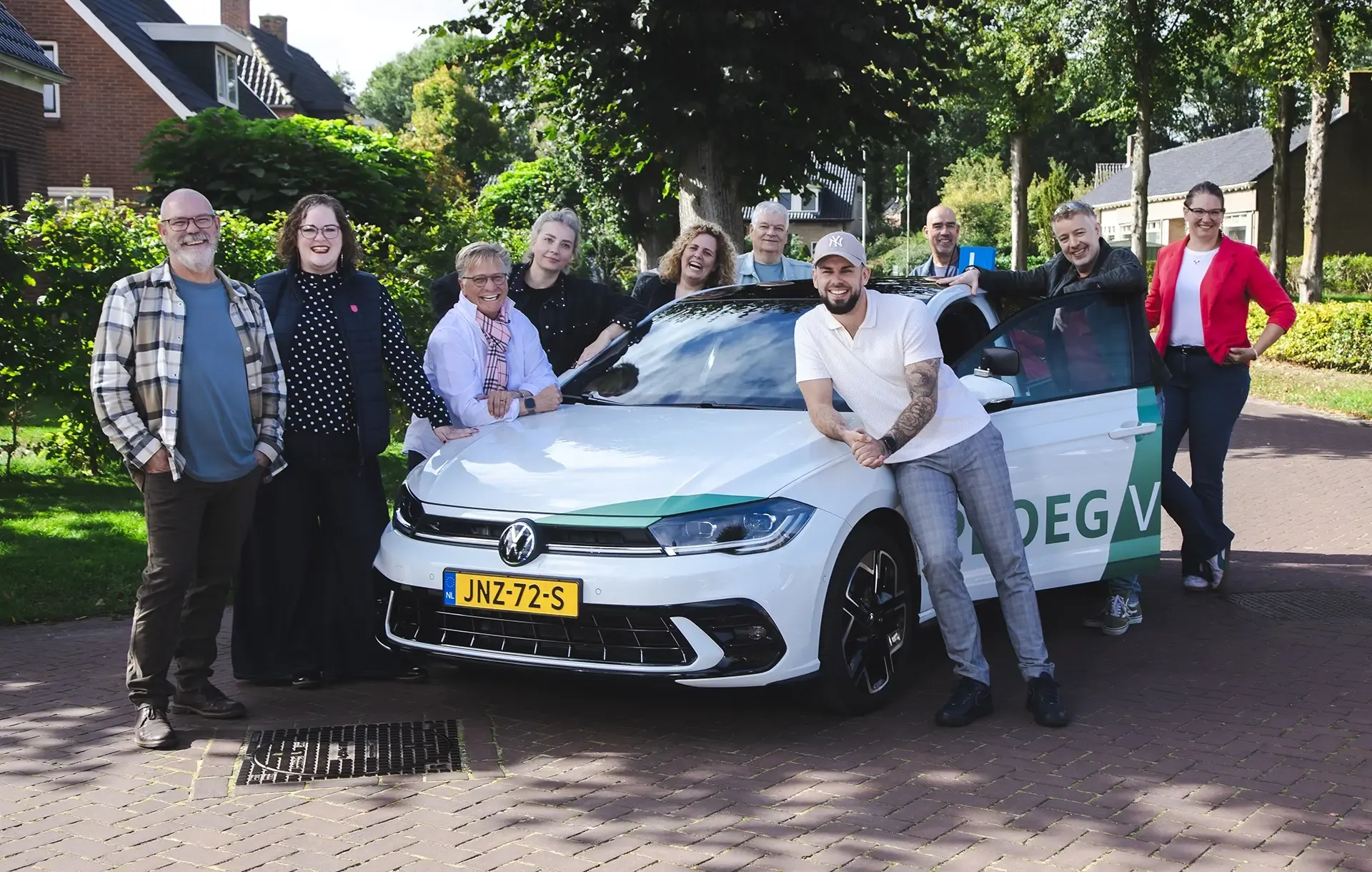 Een groep mensen poseert rond een lesauto op een zonnige dag in een woonwijk.